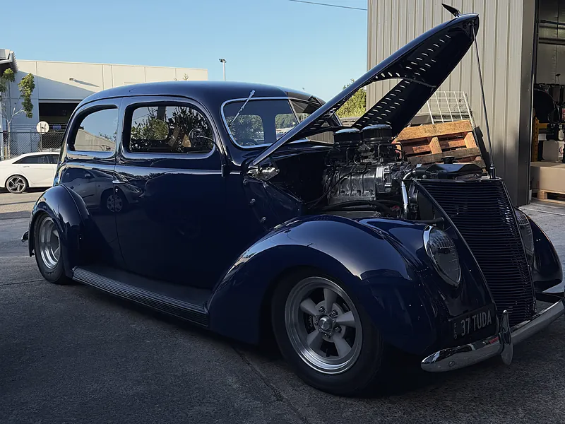 Dark blue 1937 Ford Tudor hot rod with the bonnet up outside the workshop